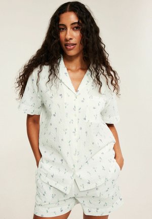 Vrouw met lang krullend haar, die een wit kortermig blouse met knopen en bijpassende korte broek met een klein bloemmotief draagt, handen in de zakken.