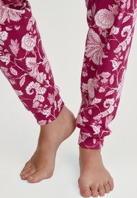 Paarse bloemenpyjamabroek met witte botanische print, rekbare katoenstof, elastische boorden en een relaxte pasvorm. Blootvoetse voeten zichtbaar.
