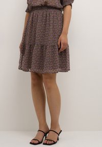 Žena v hnědých šatech střední délky s růžovým paisley vzorem a černými sandály na podpatku, stojící u prosté světlé zdi.