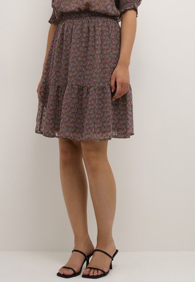 Žena v hnědých šatech střední délky s růžovým paisley vzorem a černými sandály na podpatku, stojící u prosté světlé zdi.