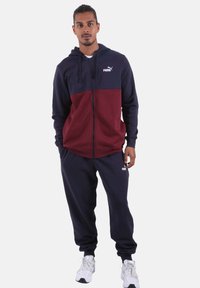 Tuta sportiva con design a blocchi di colore blu navy e bordeaux, con cappuccio, chiusura frontale con cerniera e dettagli con logo. Realizzata in tessuto morbido.