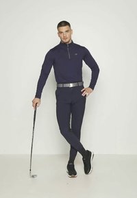 Σκούρο μπλε αθλητικό μπλούζα μακρυμάνικη με κλείσιμο quarter-zip, συνδυασμένη με εφαρμοστά παντελόνια και μαύρα αθλητικά παπούτσια, κρατώντας ένα μπαστούνι γκολφ.