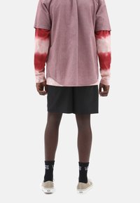 Camisa de manga corta color rojo jaspeado, superpuesta sobre una camiseta de manga larga con estampado de tie-dye. Pantalones cortos negros, calcetines negros y zapatos beige completan el conjunto.