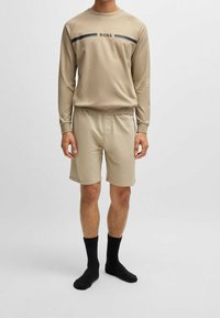 Ensemble de vêtements d'intérieur beige composé d'un sweat-shirt à manches longues et col rond avec logo noir et d'un short assorti. Des chaussettes noires complètent la tenue.