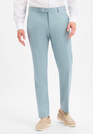 Mann trägt hellblaue Slim-Fit-Hose, weißes Hemd und beige Slipper, steht vor weißem Hintergrund.