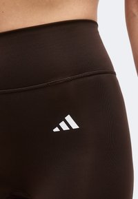 Gros plan sur un legging marron taille haute avec un logo blanc à trois bandes sur la hanche gauche, montrant un tissu lisse et les détails de la ceinture.