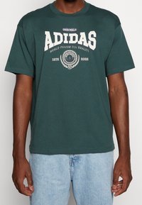 Hombre vistiendo una camiseta negra de Adidas Originals con texto y emblema blancos, combinada con unos vaqueros de mezclilla azul claro.