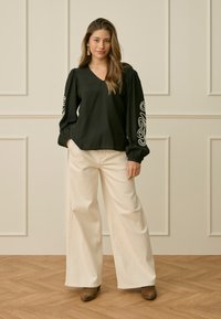 Femme debout, mains dans les poches, portant une blouse noire à manches longues avec une broderie blanche sur les manches, un pantalon large crème et des chaussures marron.