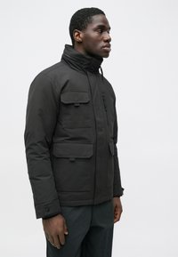 Schwarze, isolierte Jacke mit hohem Kragen, mehreren Fronttaschen und Reißverschluss. Glatte Textur und praktisches Design, geeignet für kaltes Wetter.