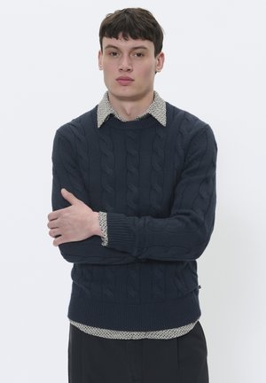 Marinefarbener Kabelstrickpullover mit geripptem Saum, über ein cremefarbenes Shirt mit Punkten getragen. Verfügt über einen Rundhalsausschnitt und lange Ärmel.