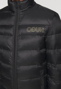 Giacca a vento nera con texture trapuntata, colletto alto e zip frontale. Presenta il logo dorato "HUGO" sul lato sinistro.