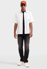 Άσπρο κοντομάνικο πουκάμισο με λεπτές κατακόρυφες ρίγες, μαύρο T-shirt από κάτω, σκούρο τζιν, άσπρα αθλητικά παπούτσια και φωτεινό πορτοκαλί καπέλο.