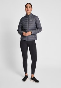 Grå puffjacka med horisontellt kviltat mönster, hög krage och dragkedja framtill, kombinerat med svarta leggings och sneakers.