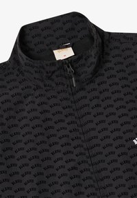 Veste noire avec un col haut, une fermeture éclair pleine et un motif "BLKVIS" répété. Fabriquée en tissu léger au toucher doux.