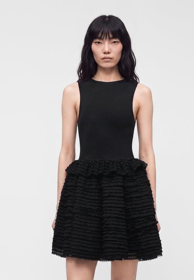 Femme aux longs cheveux noirs portant une robe noire sans manches avec une jupe texturée à volants, debout sur un fond clair uni.