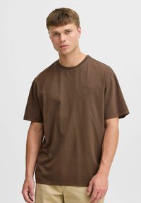Camiseta de algodón marrón con corte relajado, mangas cortas y cuello redondo. Presenta un sutil logo bordado en la parte delantera.