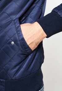 Käsi asetettuna tummansinisessä tikkitakissa, jossa on ribattu hihansuuni ja helma, hopeinen nappi näkyvissä taskussa.