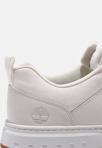 Baskets blanches à la texture lisse, présentant un logo sur le côté, un insert élastique au niveau du col, et une semelle extérieure en caoutchouc blanc avec un motif de semelle.