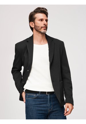 Mężczyzna o krótkich brązowych włosach i brodzie, ubrany w czarną marynarkę, białą koszulę i niebieskie dżinsy, patrzący w bok z jedną ręką w kieszeni.