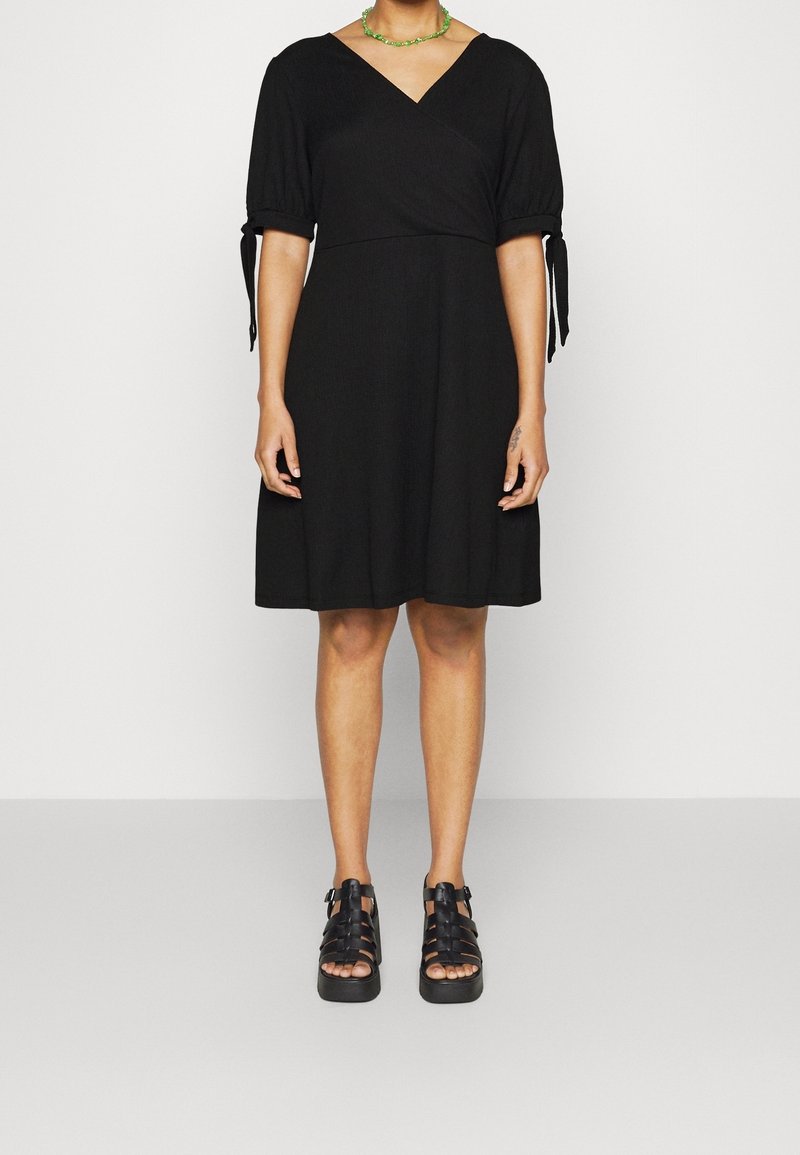 Personne portant une robe noire longueur genou avec des manches bouffantes nouées aux coudes et des sandales compensées noires.