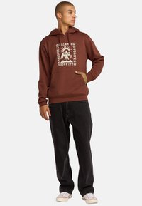 Hoodie marron avec un design graphique représentant un volcan et du texte. Fabriqué en coton, il possède une poche avant et des poignets côtelés, assorti à un jean foncé.