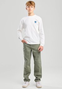 Camiseta blanca de manga larga con gráfico en el pecho; pantalones verde oliva; zapatillas blancas con detalles claros; fondo liso.