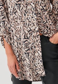 Tunic con motivo floreale in nero, crema e pesca, con una vestibilità ampia e spacchi laterali. Tessuto morbido con dettagli in bottoni e maniche arrotolate.