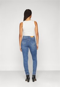 Blå denim skinny jeans med hög midja, två bakfickor och ett brunt läderpatch vid midjebandet, kombinerat med en vit tanktop.