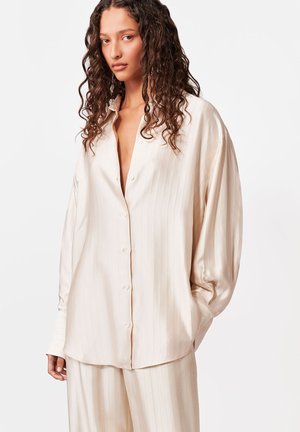 Kobieta o długich kręconych włosach, ubrana w luźną, kremową, w paski koszulę na guziki i pasujące spodnie na jednolitym białym tle.