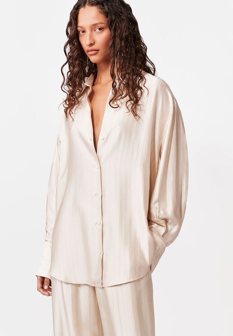 Žena s dlhými kučeravými vlasmi, oblečená v voľnej krémovej pruhovanej košeli na gombíky a zodpovedajúcich nohaviciach na jednoduchom bielom pozadí.