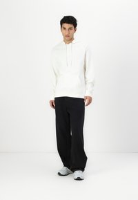 Hanorac alb din bumbac, cu buzunar frontal și sireturi, asortat cu pantaloni largi negri și adidași gri. Design curat, minimalist.