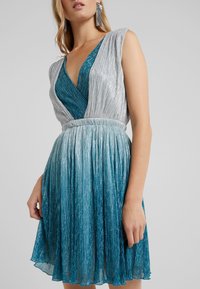 Robe présentant un design plissé aux nuances dégradées de turquoise et d'argent, avec un décolleté en V profond et des manches courtes. Tissu texturé à l'éclat métallique.