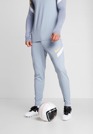 Personne en tenue de sport gris clair avec un logo Nike jaune, debout avec un pied sur un ballon de football Nike blanc et noir.