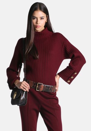Femme portant un pull côtelé bordeaux à manches larges et boutons dorés, pantalon assorti, ceinture imprimé serpent et sac à bandoulière noir.