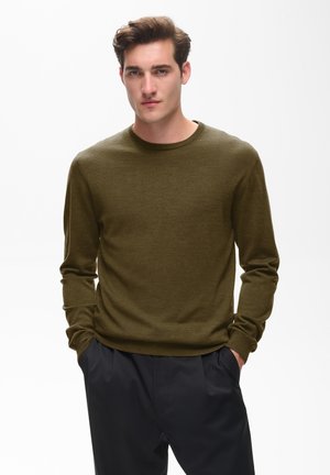 Jonge man met bruin haar, gekleed in een olijfgroene trui en donkere broek, staat met handen in de zakken tegen een effen witte achtergrond.