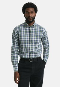 Man met baard in een groen, blauw en wit geruit button-up shirt, ingestopt in een zwarte broek met een zwarte riem, staande tegen een witte achtergrond.