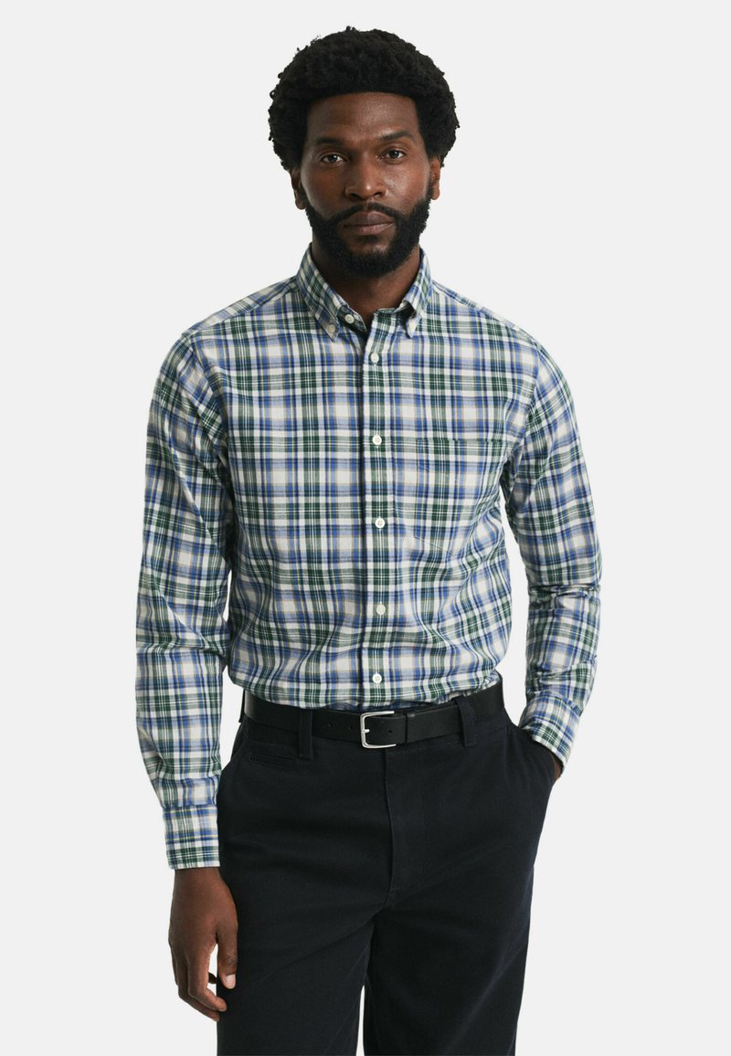 Homme avec barbe portant une chemise à carreaux verte, bleue et blanche rentrée dans un pantalon noir avec une ceinture noire, se tenant devant un fond blanc.