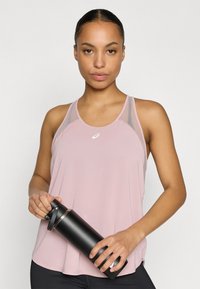 Ροζ αθλητικό τοπ ροζ με διακοσμητικά δίχτυ, με στρογγυλή λαιμόκοψη και λογότυπο. Το μοντέλο κρατά ένα κομψό μαύρο μπουκάλι νερού.