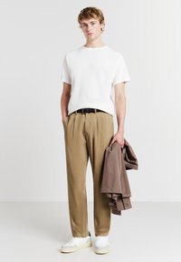 Camicia bianca a maniche corte con texture, pantaloni beige plissettati, cintura nera, tiene in mano una giacca marrone, sneakers bianche, in piedi su uno sfondo chiaro.