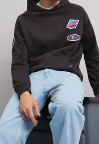 Homme portant un sweat à capuche noir avec des patchs colorés et un jean bleu clair, assis sur une commode gris foncé devant un fond uni.