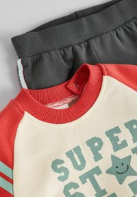 Rød og creme sweatshirt med en stjerne grafik og teal tekst, parret med mørkegrå joggere med elastisk talje. Blødt stof og ribbede manchetter.