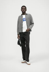 Camisa de corduroy cinza sobre uma t-shirt branca com um logótipo azul e vermelho, jeans pretos e ténis brancos com solas castanhas. Design simples e moderno.