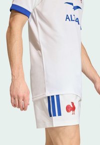 Wit shirt met blauwe accenten en Adidas-logo, gecombineerd met een witte short met een rood haan-embleem en blauwe strepen.