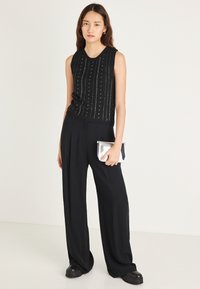 Črni brezrokavnik iz pletenine z navpičnim vzorcem, kombiniran z ohlapnimi črnimi hlačami in srebrno torbico, obuvalo črne čevlje.