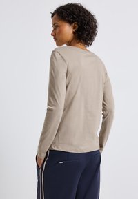 Donna con capelli ricci e scuri, che indossa un top beige a maniche lunghe e pantaloni blu navy con strisce beige, in piedi con le mani nelle tasche e rivolta di lato.