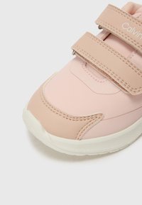 Sapatilha rosa com parte superior sintética lisa, biqueira arredondada e duas tiras de velcro. Apresenta sola de borracha e sutis detalhes de costura.