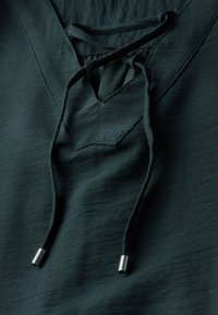Haut vert foncé avec un design en V, doté d'une fermeture lacée et de pointes en métal argenté sur les lacets. Tissu lisse et texturé.