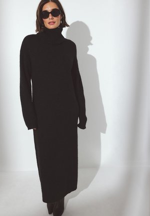 Juodas, ilgas megztinis su dideliu golfo apykakle. Minkštas audinys ir tiesi siluetas, siekiantis iki vidurinės blauzdos, dėvimas su juodais akinių nuo saulės.