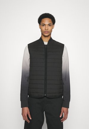 Gilet imbottito nero trapuntato con colletto alto e zip frontale, indossato sopra una camicia a maniche lunghe ombre che sfuma dal grigio al nero.