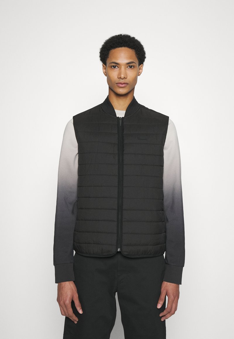Gilet imbottito nero trapuntato con colletto alto e zip frontale, indossato sopra una camicia a maniche lunghe ombre che sfuma dal grigio al nero.